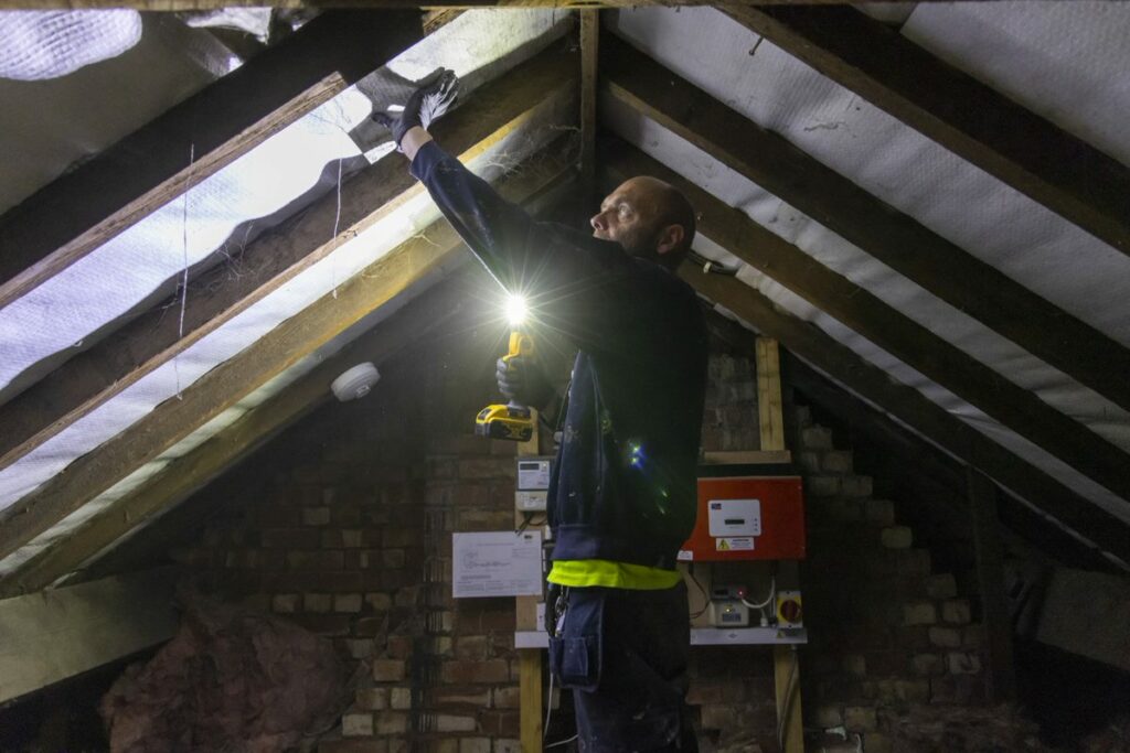 A man wearing work gloves holds a light and works on retrofitting an attic space.