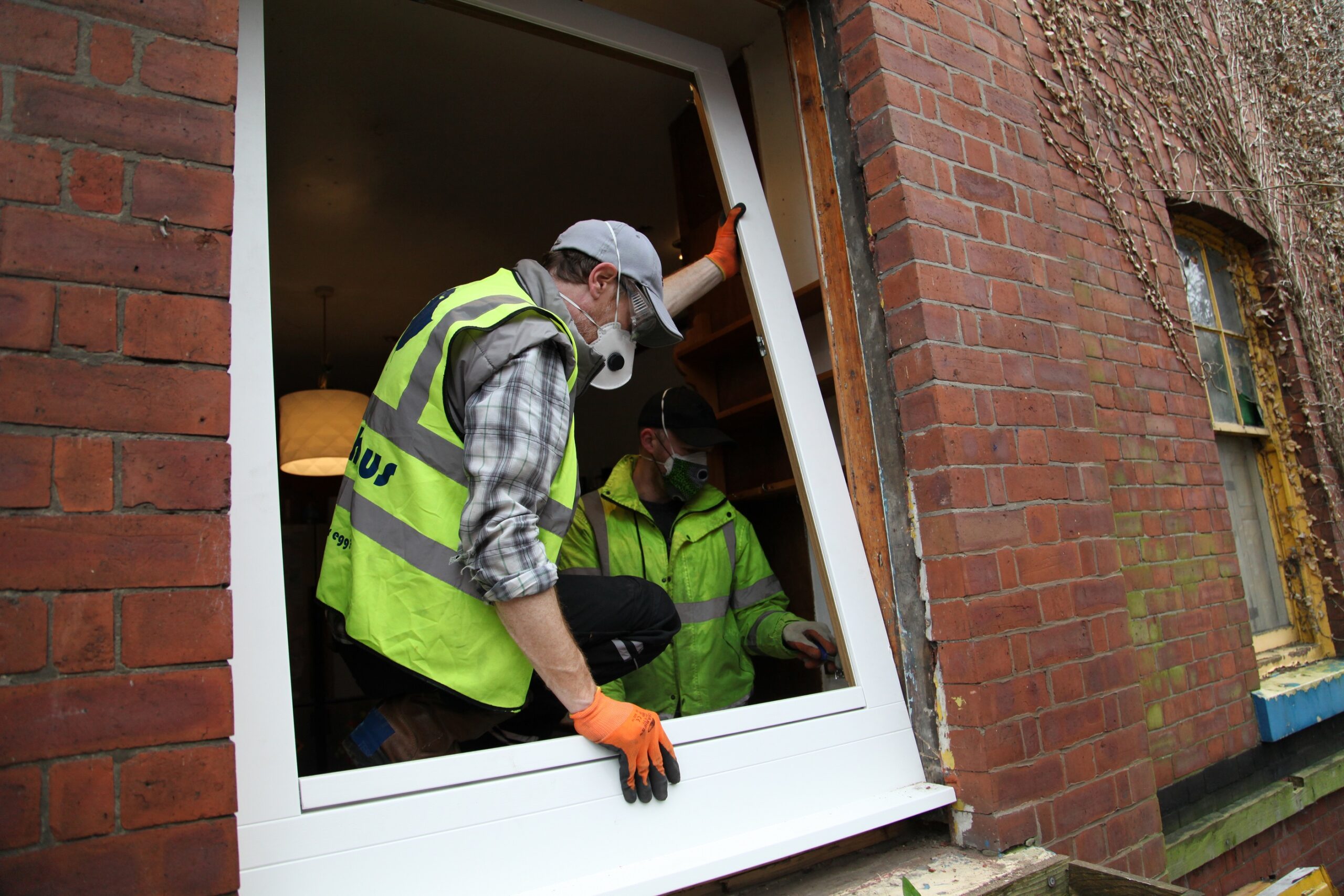 Two men in high-vis vests and work masks work on a window