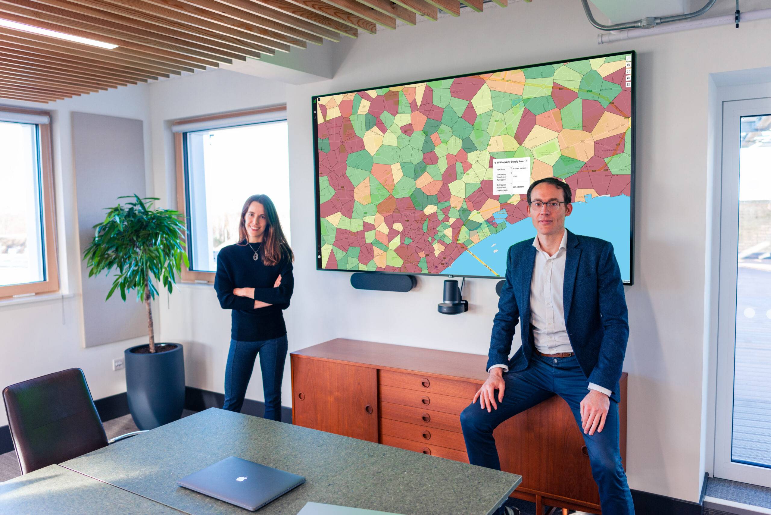 A woman (left) and man (right) in professional attire stand in a conference room in front of a large screen displaying a colorful geospatial grid system map.