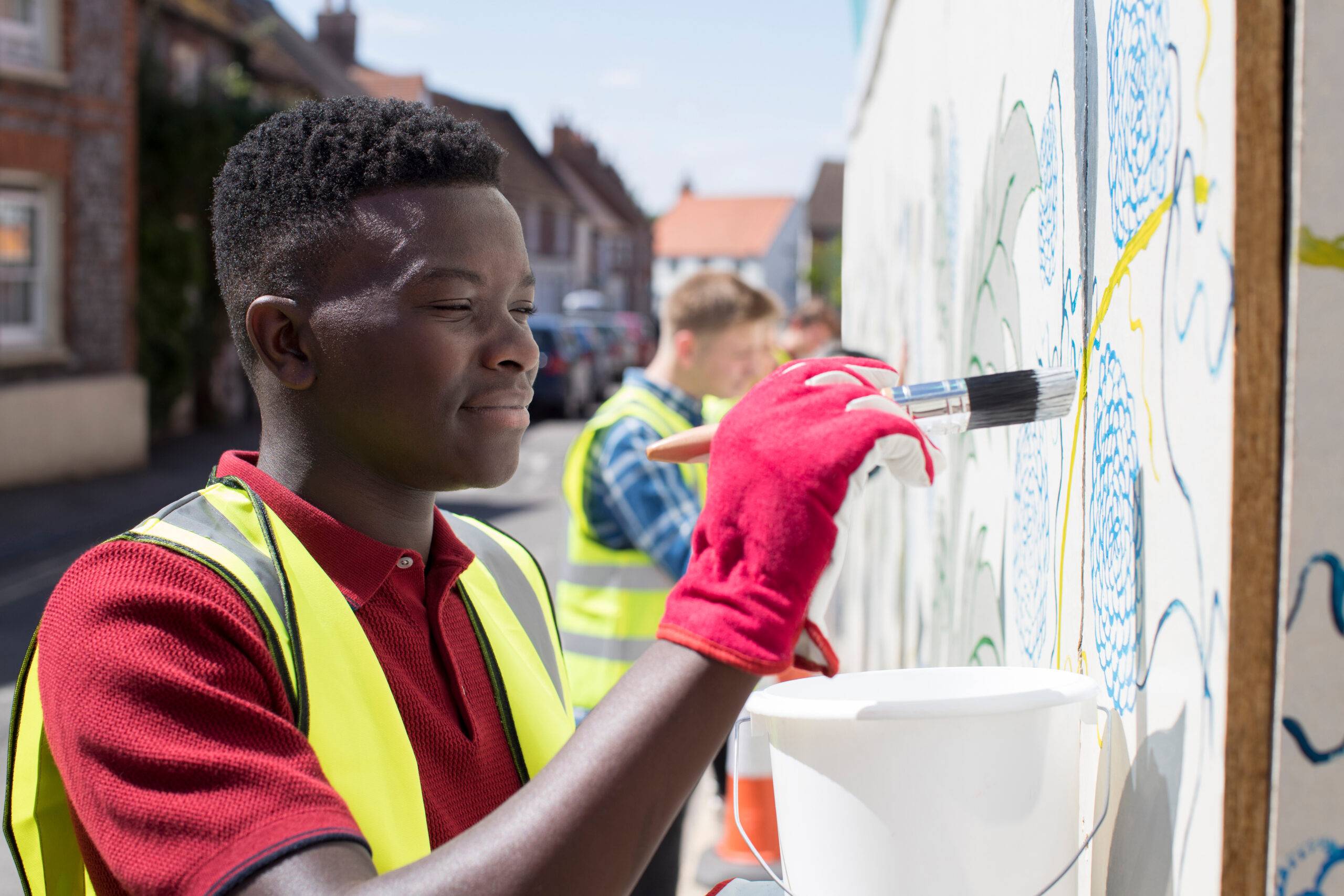 Young man wears work gloves and high-vis vest while painting a wall.