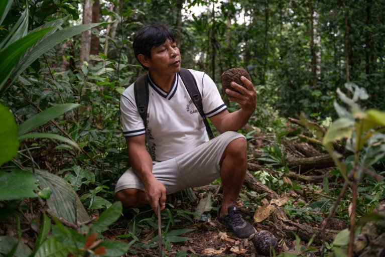 A man in a forest holds a coconut and a farming tool