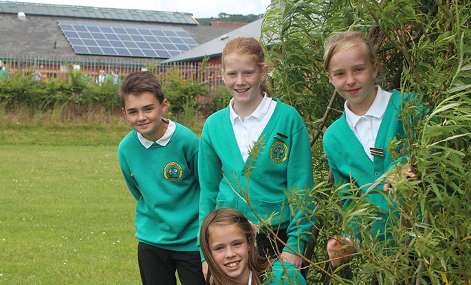 4 school children stand in their grassy school field smiling at the camera. There are solar panels on the school building's roof visible behind them.