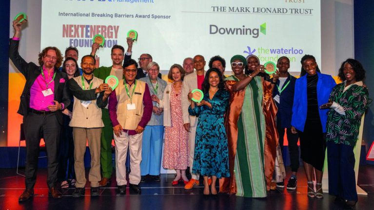The 2025 Ashden Award Winners are stood on stage cheering whilst holding their awards.