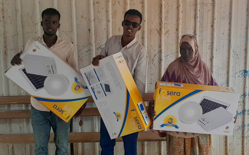 Three community members are holding up solar powered lights that have been given to them by Muruqmaal Energy and Electric Cooperative.