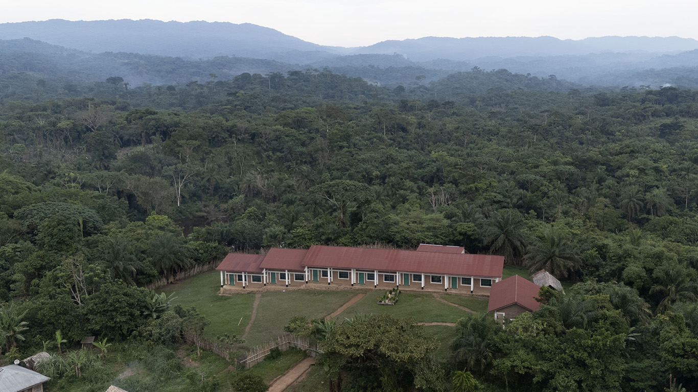 Aerial view of a long, single-story building with a red roof and white pillars, surrounded by dense tropical rainforest. The building sits in a small clearing with dirt paths and minimal development. Green trees dominate the landscape, stretching to misty hills and mountains in the background under a cloudy sky.