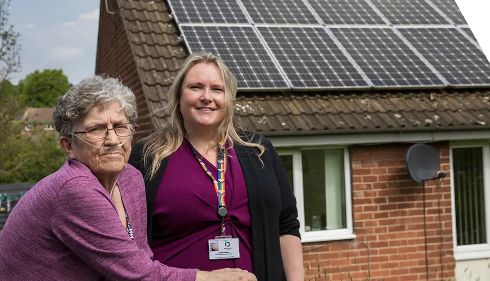 An elderly lady, with respiratory equipment to aid with her breathing, stands next to a local council member who has helped her to install solar panels on her roof.