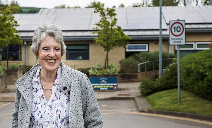 A woman smiles whilst standing in front of a school that has had solar panels installed on its roof.